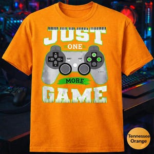 May include: Orange t-shirt with a grey and green video game controller design. The text "JUST ONE MORE GAME" is printed above and below the controller in white and green.