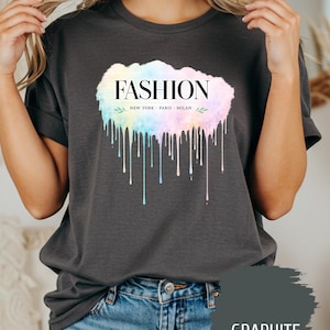 May include: A graphite-coloured t-shirt featuring the word "FASHION" in black, with "NEW YORK PARIS MILAN" below. The word "FASHION" is set against a watercolour cloud in pastel shades with dripping paint effects.