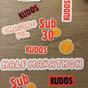 May include: A selection of white stickers with red and black text on a wooden surface. The stickers display phrases such as "Powered by Pastries," "Kudos," "Half Marathon," and "Local Legend." Some stickers include medal graphics.