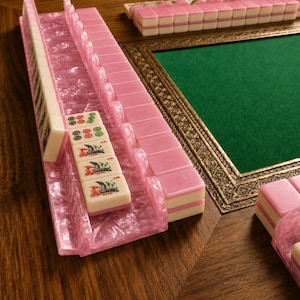 Op de afbeelding: Een roze en witte Mahjong-set met een roze rek, gerangschikt op een houten oppervlak. De tegels zijn wit met roze accenten en symbolen. Op de achtergrond een groen viltgevoerd tafel met een decoratief gouden frame.