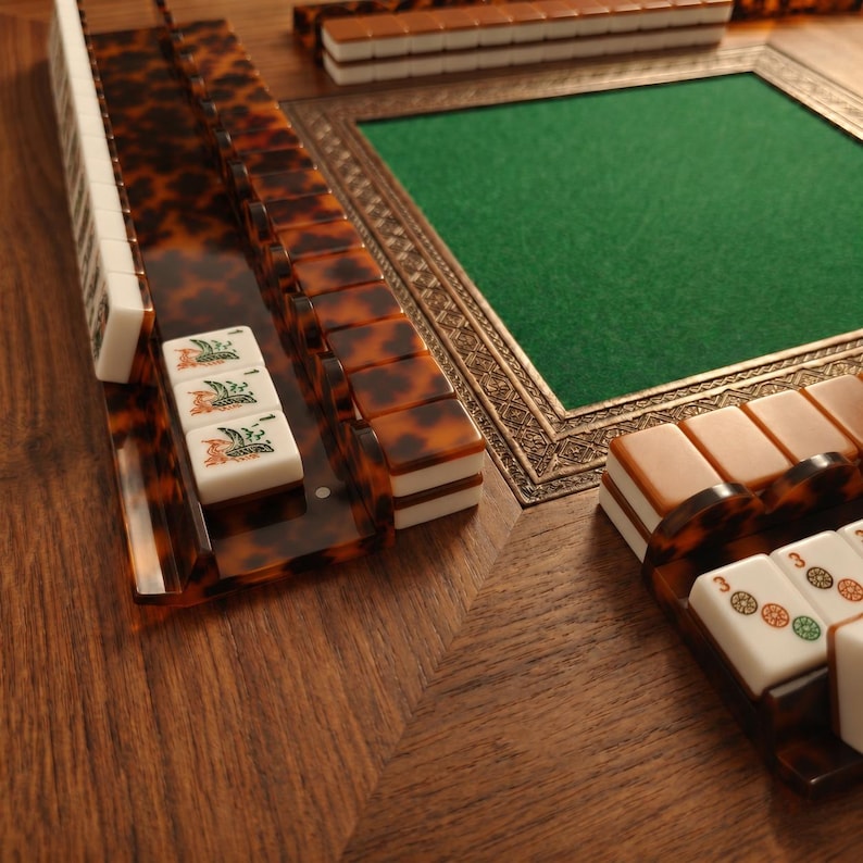 Pode incluir: Um close-up de um conjunto de Mahjong com suportes e pe&ccedil;as com padr&atilde;o de tartaruga. O conjunto inclui uma superf&iacute;cie de jogo de feltro verde emoldurada em ouro. As pe&ccedil;as s&atilde;o brancas com s&iacute;mbolos e n&uacute;meros detalhados.