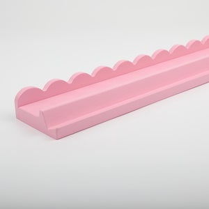 Puede incluir: Una estanter&iacute;a rosa claro con borde festoneado. La estanter&iacute;a es rectangular con un borde frontal elevado y un borde superior festoneado. La estanter&iacute;a est&aacute; pintada de un color rosa suave y liso.
