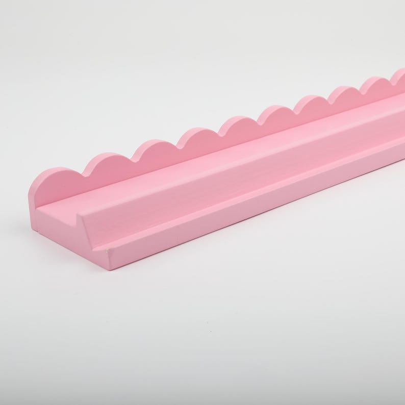 Puede incluir: Una estanter&iacute;a rosa claro con borde festoneado. La estanter&iacute;a es rectangular con un borde frontal elevado y un borde superior festoneado. La estanter&iacute;a est&aacute; pintada de un color rosa suave y liso.
