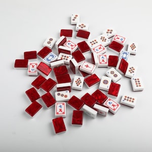May include: A collection of rectangular Mahjong tiles scattered on a surface. The tiles feature white faces with various colorful Chinese characters, numbers, and circular patterns, while the backs are a deep, marbled translucent red color.