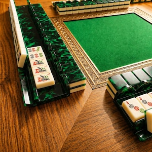 May include: A Mahjong set with green and black tiles and racks, displayed on a wooden table. The set features a green felt playing surface with a decorative gold frame. The white tiles display various symbols and characters, ready for play.