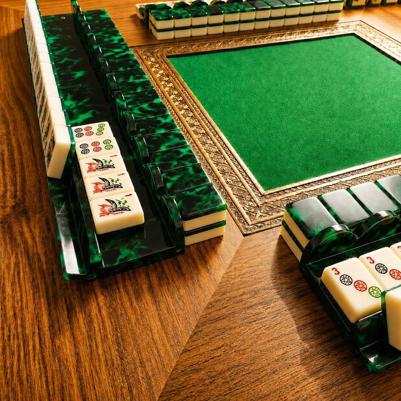 May include: A Mahjong set with green and black tiles and racks, displayed on a wooden table. The set features a green felt playing surface with a decorative gold frame. The white tiles display various symbols and characters, ready for play.