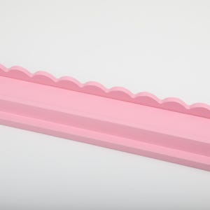 Puede incluir: Estante de madera rosa claro con borde festoneado decorativo. El estante tiene una base plana y un borde frontal elevado. El estante est&aacute; pintado de un color rosa suave y s&oacute;lido y est&aacute; sobre un fondo blanco.