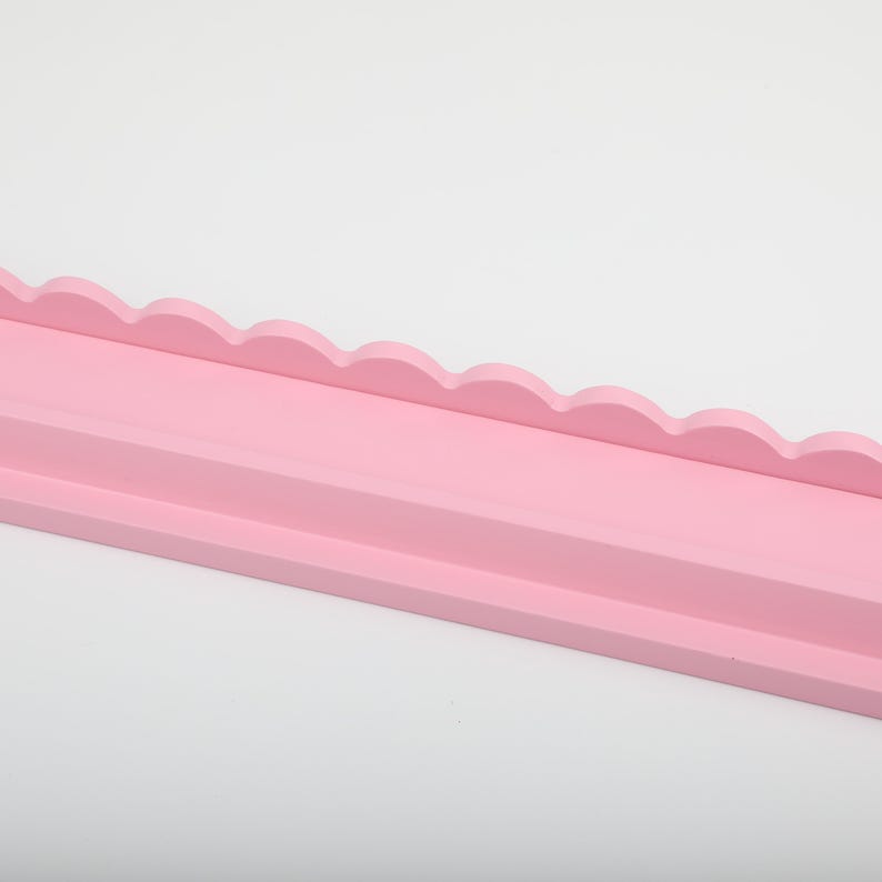 Puede incluir: Estante de madera rosa claro con borde festoneado decorativo. El estante tiene una base plana y un borde frontal elevado. El estante est&aacute; pintado de un color rosa suave y s&oacute;lido y est&aacute; sobre un fondo blanco.