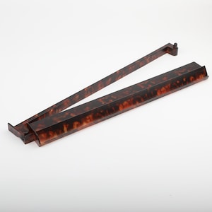 May include: A pair of vintage-style, tortoise-shell patterned serving tongs. The tongs are made of a dark brown material with lighter brown and amber marbling. The tongs are open and angled, with a rectangular base and a long, slender handle.