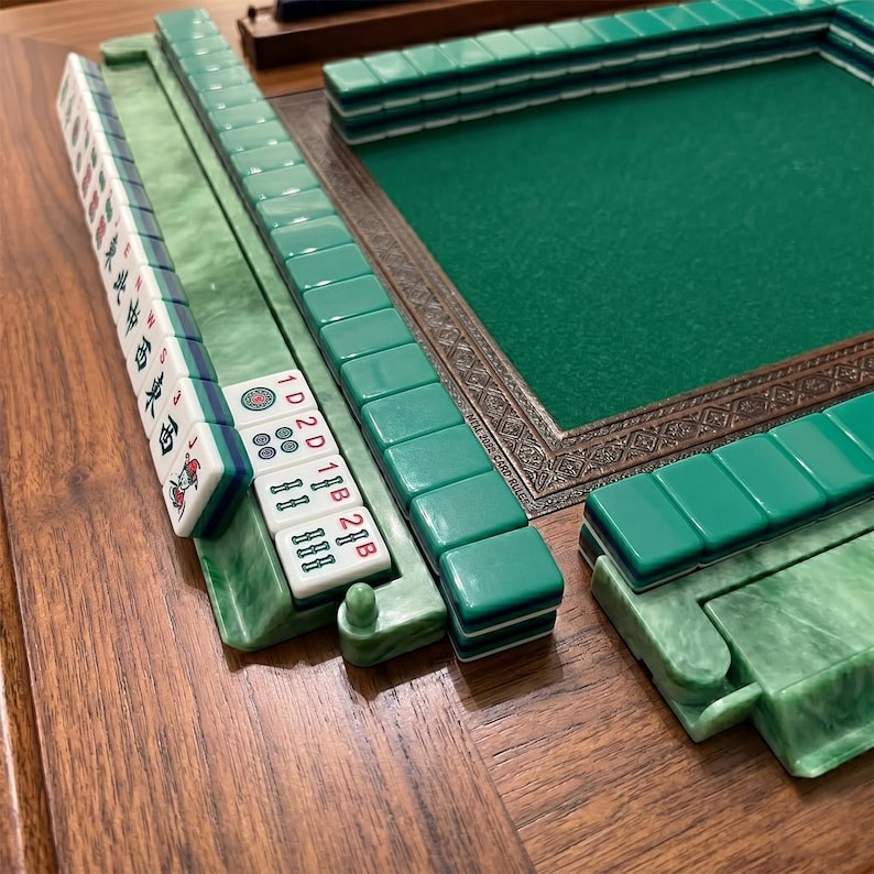 May include: A close-up of a Mahjong set with green and white tiles. The tiles are arranged on a wooden surface, with a green felt playing surface in the background. The set includes racks and a decorative border.