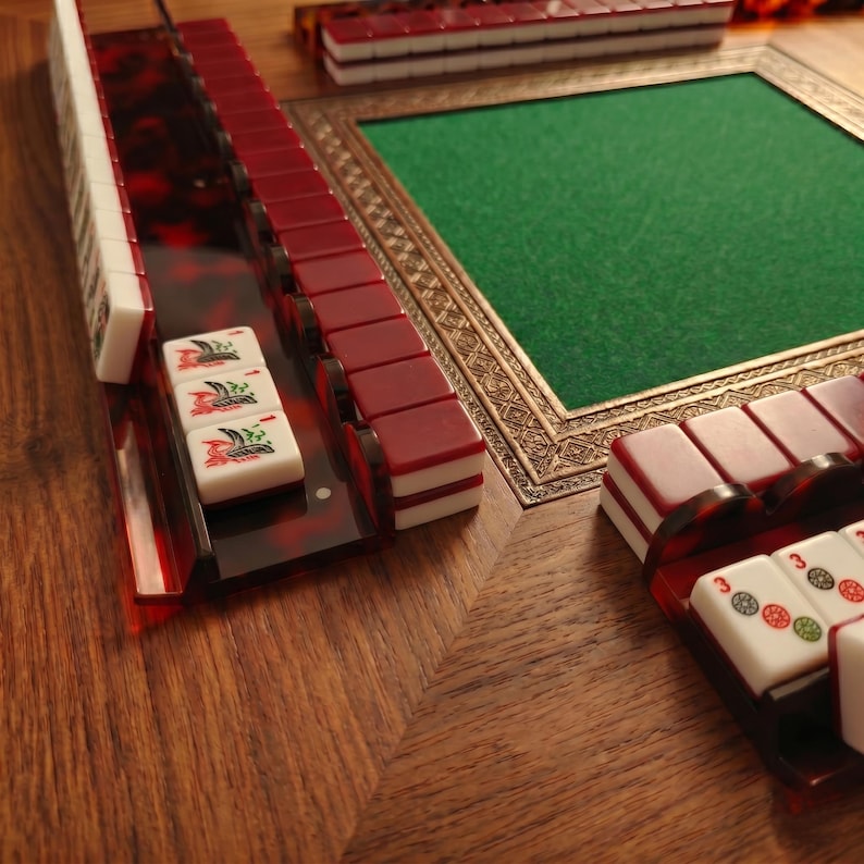 Puede incluir: Primer plano de un juego de Mahjong con fichas rojas y blancas, dispuestas sobre una superficie de madera. Las fichas est&aacute;n en un soporte rojo y marr&oacute;n. Un tapete de juego verde forrado de fieltro est&aacute; en el fondo.