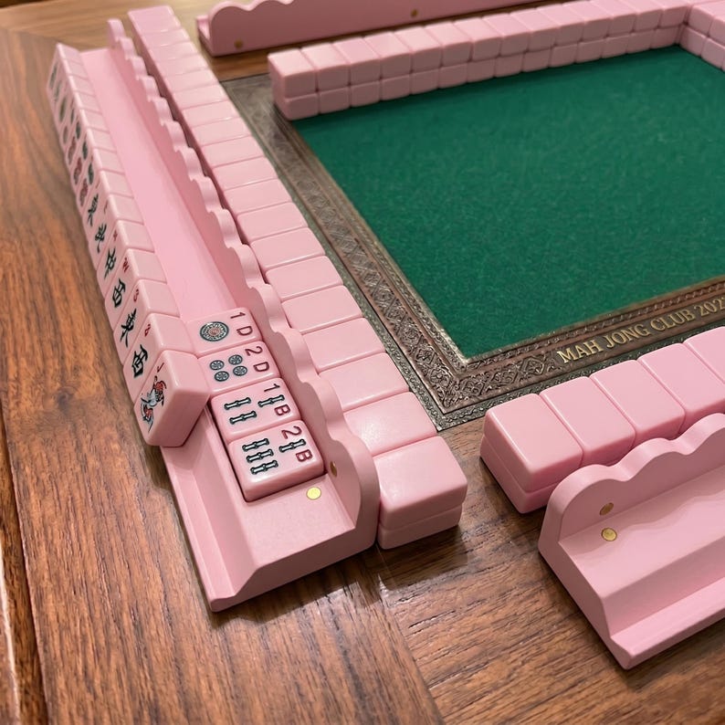 Puede incluir: Un juego de Mahjong rosa con fichas y estantes a juego. El juego incluye una superficie de juego verde con un borde decorativo. El texto "MAH JONG CLUB 2023" es visible en la superficie de juego.