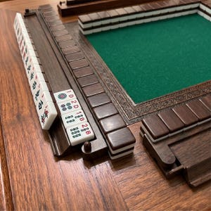 Könnte beinhalten: Ein Mahjong-Set aus Holz mit einer grünen Spielfläche. Das Set enthält braune Holzgestelle, weiße Spielsteine mit roten und schwarzen Markierungen und einen dekorativen Rahmen. Die Spielsteine sind auf einer Holzoberfläche angeordnet.