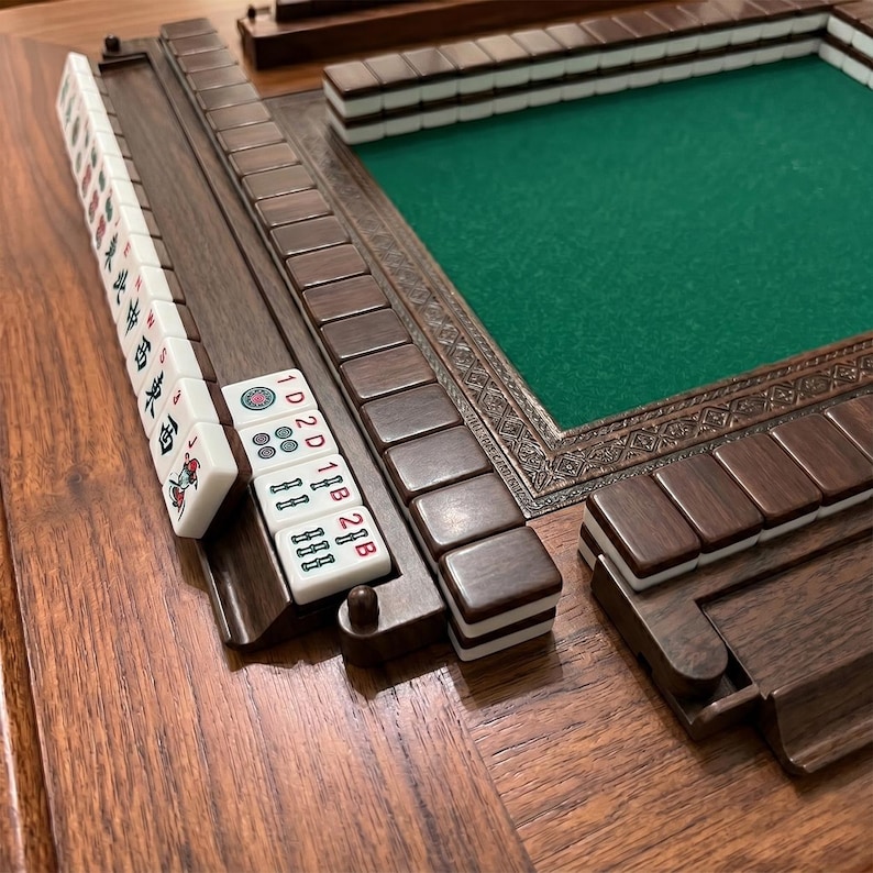 K&ouml;nnte beinhalten: Ein Mahjong-Set aus Holz mit einer gr&uuml;nen Spielfl&auml;che. Das Set enth&auml;lt braune Holzgestelle, wei&szlig;e Spielsteine mit roten und schwarzen Markierungen und einen dekorativen Rahmen. Die Spielsteine sind auf einer Holzoberfl&auml;che angeordnet.