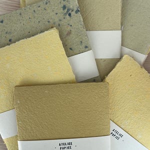Op de afbeelding: Een verzameling rechthoekige handgeschepte vellen papier in diverse tinten geel, beige en groen, met geschepte randen en gestructureerde oppervlakken. Elke stapel is vastgezet met een witte papieren band met de tekst ATELJEE PAPJEE.