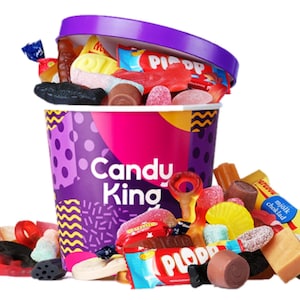 Puede incluir: Un cubo morado y rosa rebosante de caramelos de colores. El cubo tiene la inscripción "Candy King". Varios caramelos, incluyendo chocolates y gominolas, están esparcidos.