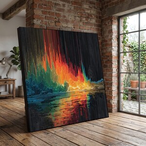 Op de afbeelding: Groot, vierkant abstract kunstwerk met een zwart frame. Het kunstwerk toont een kleurrijk, gepixeld landschap met levendige tinten oranje, geel, groen en blauw, weerspiegeld in een donker, waterachtig oppervlak. Het kunstwerk leunt tegen een bakstenen muur.