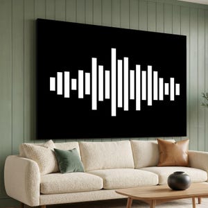 Op de afbeelding: Een groot zwart canvas wandkunstwerk met een wit geluidsgolfontwerp. De geluidsgolf bestaat uit verticale balken van verschillende hoogtes, die geluid visueel weergeven. Het kunstwerk wordt boven een beige bank weergegeven.