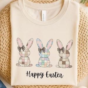 Könnte beinhalten: Cremefarbenes T-Shirt mit drei Osterhasen-Designs mit Patchworkmustern in Pastellfarben. Jeder Hase hat eine Schleife und die Worte "Happy Easter" sind darunter gedruckt.