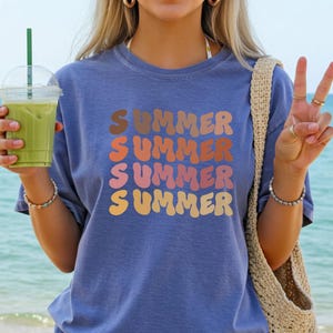 May include: Blue t-shirt with the word "SUMMER" in stacked, retro-style lettering in shades of brown, orange, pink, and yellow. The shirt is worn on a beach with a drink in hand. A beige woven bag is on the shoulder.