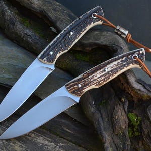 Peut inclure: Deux couteaux à lame fixe avec des lames en acier argenté et des manches en bois de cerf brun texturé. Chaque manche est fixé par deux rivets métalliques et comporte une lanière en cuir brun avec une perle en bois de cerf.