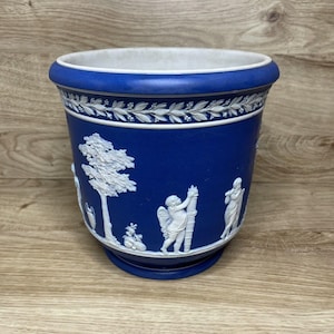 May include: A deep blue cylindrical planter featuring white relief ornamentation. The design includes a laurel leaf border at the top and a continuous scene of white figures, trees, and foliage wrapped around the exterior of the vessel.