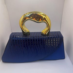 May include: A blue handbag with a gold-colored handle. The bag has a textured, crocodile-like pattern on the front and a solid blue side panel. The handle is a curved, arched design.