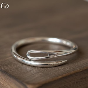 Può includere: Un anello color argento a forma di ago, con una fascia curva e un design a occhiello. L'anello è presentato su una superficie di legno scuro. Il logo RingLabCo è visibile in alto a sinistra.