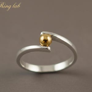 Könnte beinhalten: Ein silberner Ring mit einer goldfarbenen Kugel in der Mitte. Das Ringband umschließt den Finger, wobei die Kugel zwischen den beiden Enden gehalten wird. Der Text "Ring lab" ist oben links zu sehen.