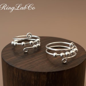 Peut inclure: Deux bagues argentées avec un design en spirale et de petites perles rondes. Les bagues sont présentées sur une surface en bois brun foncé. Le texte "Ring Lab Co" est dans le coin supérieur gauche.