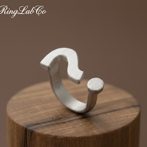 May include: A silver ring shaped like a question mark, with a small circular detail. The ring is presented on a wooden block. The brand name "Ring Lab Co" is visible in the upper left corner.