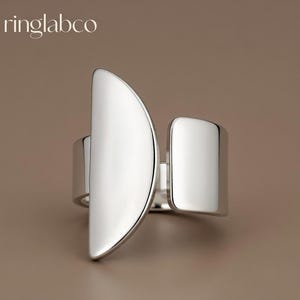 Puede incluir: Un moderno anillo de plata con un diseño único. El anillo presenta una gran forma de media luna en un lado y una forma rectangular en el otro. El anillo está muy pulido y refleja la luz. La marca "ringlabco" está en la esquina superior izquierda.