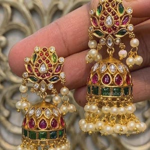 Peut inclure: Une paire de boucles d'oreilles jhumka dorées avec un clou en forme de lotus orné de pierres rouges et vertes, de cristaux clairs et de perles blanches pendantes. La base en cloche présente des pierres rouges et vertes et des perles.