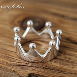 May include: A silver crown ring with six rounded points, each topped with a small sphere. The ring is set against a wooden background. The word "ringlabco" is in the upper left corner.