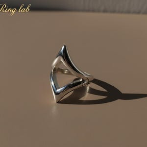 May include: A silver ring with a distinctive open design. The ring has a V-shaped element, creating an open space in the centre. The ring is set against a neutral background, with the text "Ring lab" in the upper left corner.