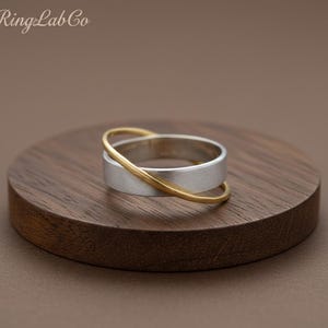 May include: A silver ring with a brushed finish, crossed by a thin gold band. The ring is displayed on a dark wooden surface. The text "RingLab Co" is in the upper left corner.