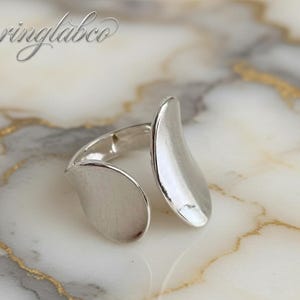 Puede incluir: Un anillo plateado con un diseño abierto. El anillo presenta dos elementos curvos en forma de hoja que se envuelven alrededor del dedo. El anillo está sobre un fondo de mármol, con el texto "ringlabco" en la esquina superior izquierda.