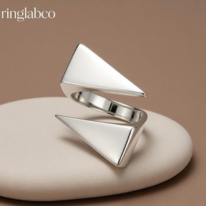 Può includere: Un anello color argento dal design geometrico. L'anello presenta due forme triangolari collegate da una fascia. È esposto su una superficie arrotondata di colore chiaro. Il nome del marchio "ringlabco" è visibile.