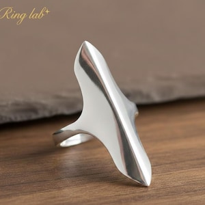 Puede incluir: Un anillo plateado con un diseño largo y puntiagudo. El anillo tiene una superficie lisa y pulida y una forma única y alargada. El anillo está sobre una superficie de madera. El texto "Ring lab" es visible.