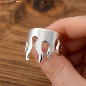 Pode incluir: Um anel prateado com um design de chama. O anel é segurado por uma mão, mostrando a forma única da joia. O design do anel apresenta recortes curvos em forma de chama ao redor da banda.