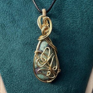 May include: A handcrafted pendant featuring a teardrop-shaped stone wrapped in gold-colored wire. The stone has a mottled green and purple pattern. The wirework forms decorative swirls and a bail for a black cord necklace. The pendant is displayed against a teal background.