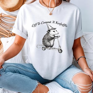 Könnte beinhalten: Weißes T-Shirt mit dem Text "Off To Commit A Kerfuffle" und einer Schwarz-Weiß-Illustration eines Opossums auf einem Roller mit Hexenhut. Ein lässiges Unisex-T-Shirt.