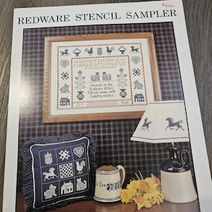 May include: A vintage "Redware Stencil Sampler" pattern book with a framed cross-stitch design. The sampler features a variety of country-themed motifs, including animals and houses. A blue pillow, mug, and lamp with horse designs are also visible.