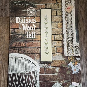 May include: Vintage cross-stitch pattern titled "Daisies Won't Tell" with a floral design. The cover features a brick wall background, a hanging cross-stitch piece, and a wicker chair. A price tag is visible.