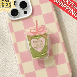 May include: A phone case with a pink and white chequered pattern. A clear, acrylic phone grip in the shape of a matcha drink is attached. The grip has a pink bow and the words "Matcha Matcha Matcha" inside a heart.