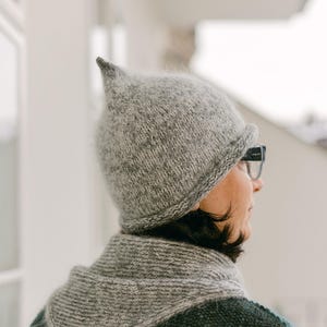 May include: A gray knit hat with a pointed top and a matching scarf. The hat and scarf are made of a soft, textured yarn. The person is wearing glasses and a patterned sweater.