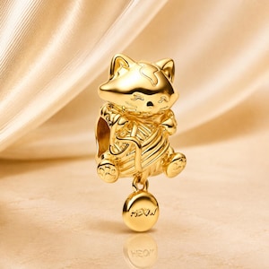 Può includere: Un charm dorato raffigurante un gatto che tiene in mano un gomitolo di lana. Il charm ha un ciondolo rotondo con la scritta "MEOW". Il gatto è seduto e ha una faccia sorridente. Il charm è realizzato in un materiale lucido e riflettente.