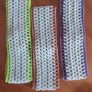 May include: Three rectangular crochet pieces in a row. Each piece is a light gray color with a colored border. The borders are lime green, peach, and purple. The crochet has a grid-like pattern.