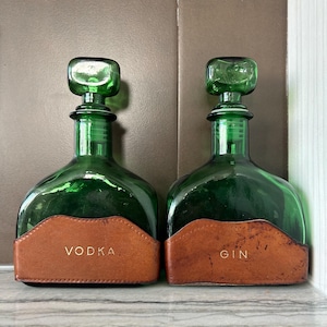 May include: Two green glass decanters with square stoppers. Each decanter has a brown leather label. One label reads "VODKA" and the other reads "GIN". The decanters are displayed on a white surface.