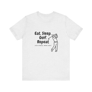 Op de afbeelding: Lichtgrijs t-shirt met de tekst "Eat. Sleep. Golf. Repeat" en een zwarte illustratie van een golfer. De zin "LESS STRESS, MORE GOLF" staat eronder, voor golfliefhebbers.